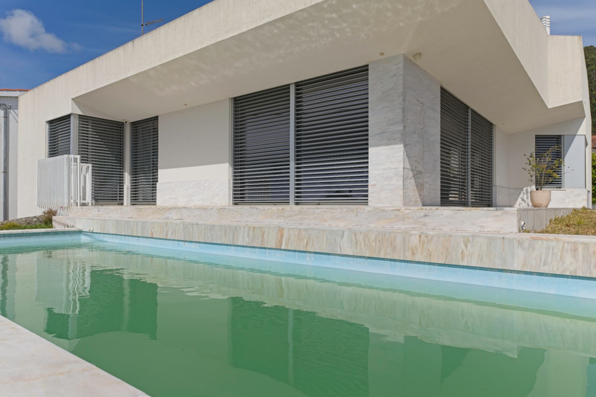 Piscina com bordadura e revestimento em pedra natural clara junto à fachada de uma habitação.