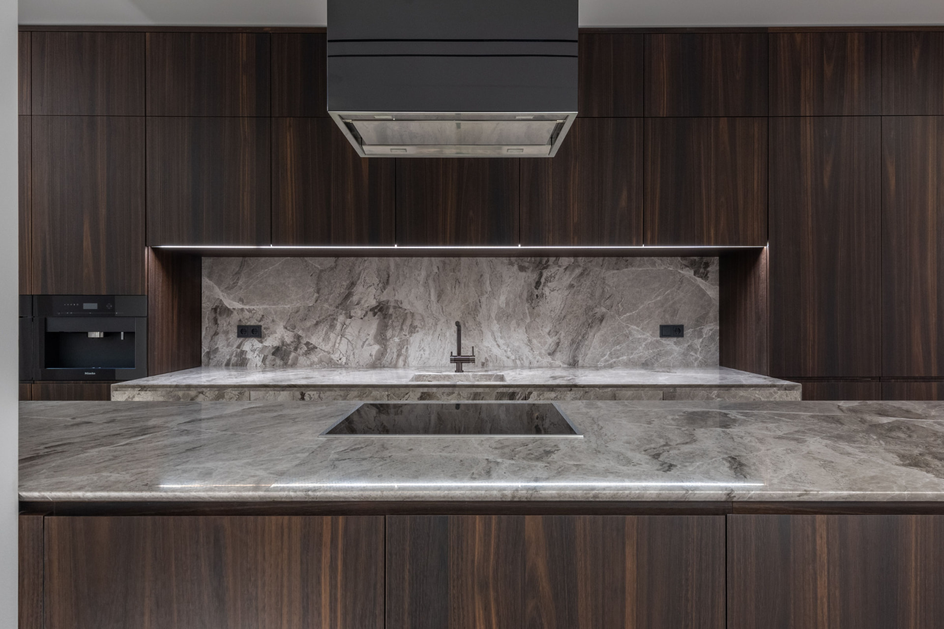 Cozinha contemporânea com bancada e painel em pedra natural cinzenta e mobiliário em madeira escura.