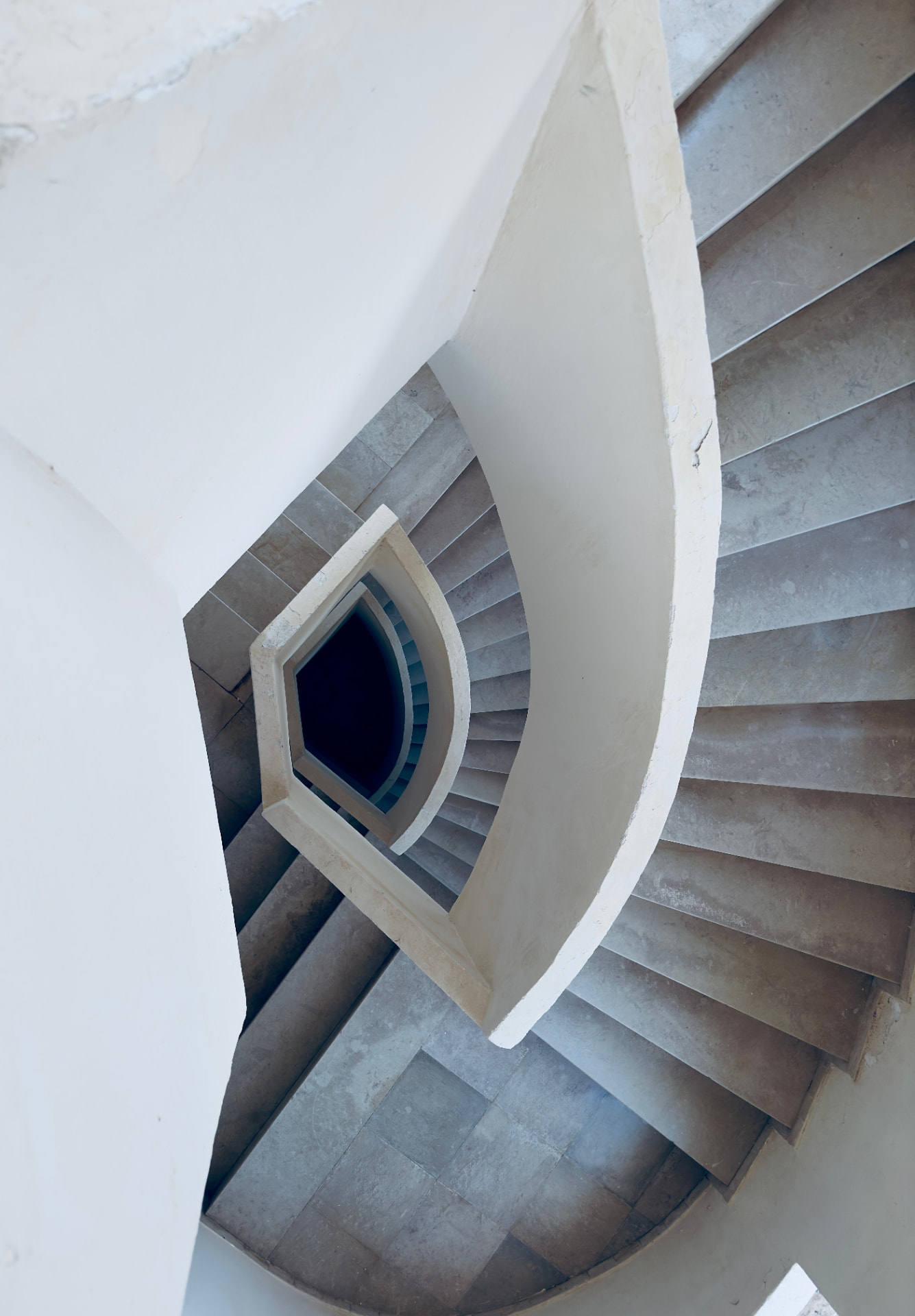 Escada espiral em pedra clara vista de cima, com degraus curvos em torno do vazio central.