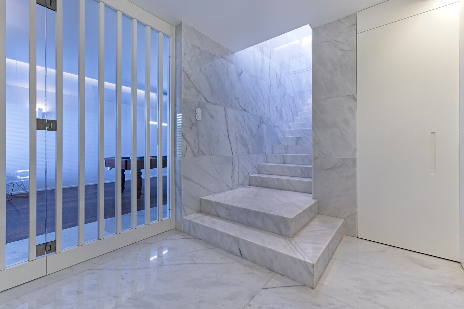 Escada e paredes interiores revestidas a mármore branco, junto a uma porta em vidro com caixilhos.