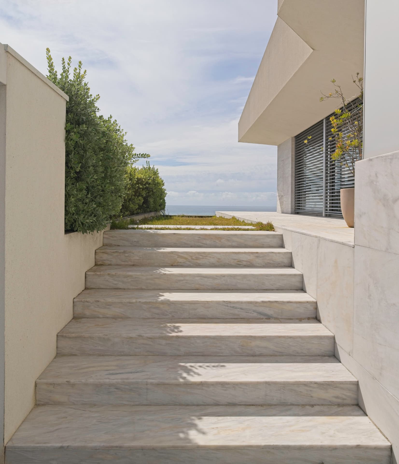 Escadas exteriores em mármore claro a descer para um jardim, junto a uma casa contemporânea com vista de mar.